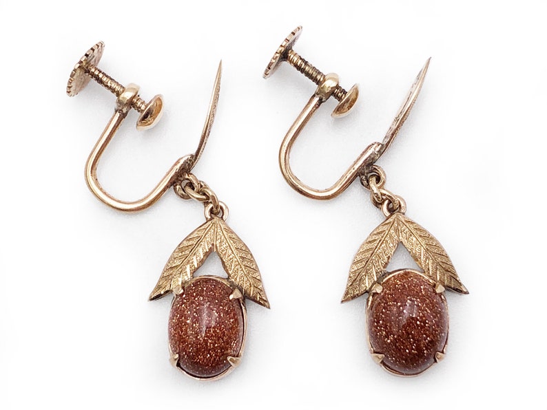 May include: A pair of gold tone clip-on earrings with brown stone accents. Each earring features a teardrop-shaped stone with a gold tone leaf design.