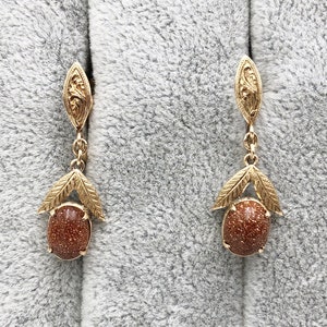 May include: A pair of gold earrings with brown gemstone drops. The earrings feature a delicate floral design and are suspended from a small, ornate loop.