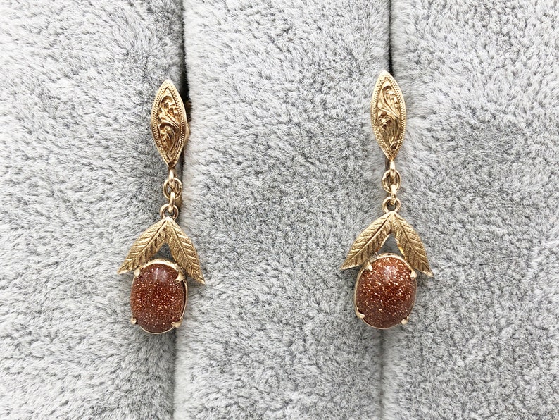 May include: A pair of gold earrings with brown gemstone drops. The earrings feature a delicate floral design and are suspended from a small, ornate loop.