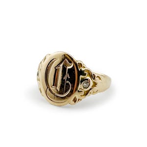 May include: A gold signet ring with an oval face featuring the letter "C" in an ornate script. The ring has a small diamond accent and decorative filigree detailing. The ring is set against a white background.