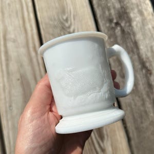 May include: White milk glass mug with a handle and a pedestal base. The mug features a raised relief design of a rabbit. The mug is held in a hand against a wooden background.