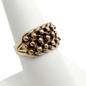 K&ouml;nnte beinhalten: Ein goldener Ring mit einem strukturierten Design aus kleinen, runden Perlen. Der Ring befindet sich auf einem wei&szlig;en Pr&auml;sentationsst&auml;nder.
