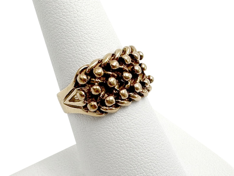 K&ouml;nnte beinhalten: Ein goldener Ring mit einem strukturierten Design aus kleinen, runden Perlen. Der Ring befindet sich auf einem wei&szlig;en Pr&auml;sentationsst&auml;nder.