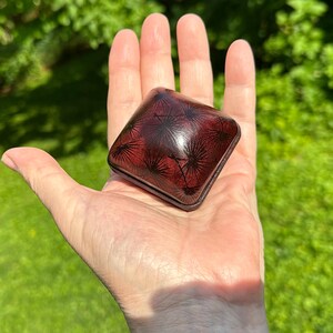 Pode incluir: Uma pequena caixa quadrada de cor vermelho-escura com cantos arredondados e acabamento brilhante. A tampa apresenta um padrão preto repetitivo de sementes de dente-de-leão. A caixa é compacta, desenhada para caber na palma da mão.