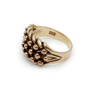 K&ouml;nnte beinhalten: Ein goldener Ring mit einem strukturierten Design aus einer Ansammlung kleiner Kugeln. Der Ring hat eine schlichte Band und ist innen mit den Buchstaben "CU" gekennzeichnet.