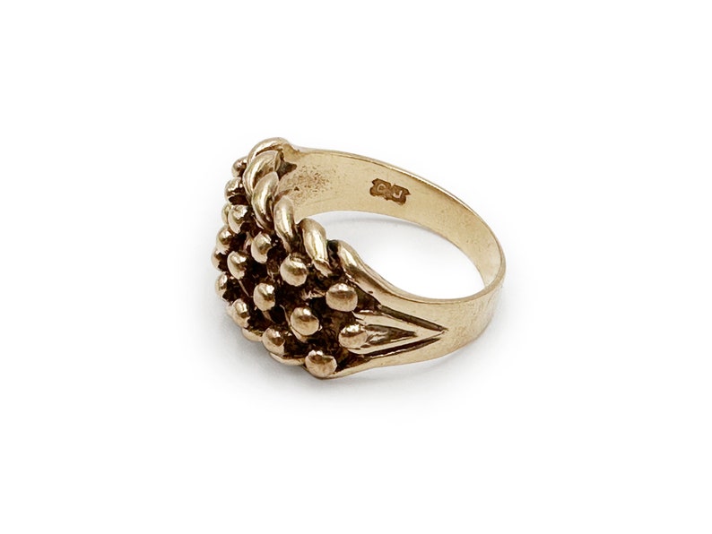 K&ouml;nnte beinhalten: Ein goldener Ring mit einem strukturierten Design aus einer Ansammlung kleiner Kugeln. Der Ring hat eine schlichte Band und ist innen mit den Buchstaben "CU" gekennzeichnet.