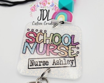 May include: A white glitter badge holder with a rainbow design and the text "School Nurse: Nurse Ashley". The badge holder is attached to a black lanyard with colorful silicone beads.