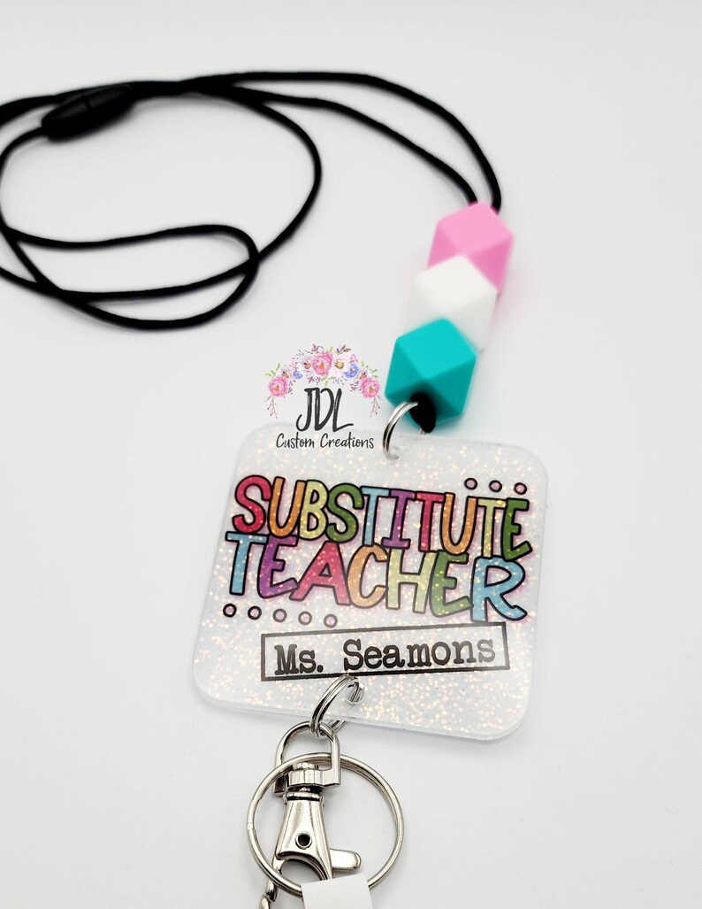 May include: A white, glittery badge holder with the text "Substitute Teacher Ms. Seamons" in rainbow colors. The badge holder is attached to a black lanyard with a turquoise, white, and pink silicone bead charm. A silver key ring is attached to the bottom of the badge holder.