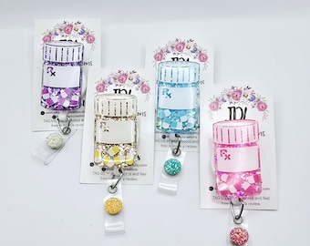 May include: Four colorful pill bottle-shaped retractable badge holders with glitter and floral designs. Each holder has a  "Rx" symbol and a white label. The holders are attached to a clear retractable badge reel with a decorative charm.