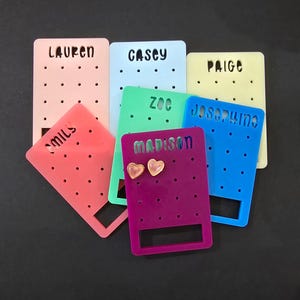 May include: A collection of colorful acrylic earring display cards in pink, blue, green, purple, and yellow. Each card has a name cut out at the top and small holes for earrings. The purple card displays heart-shaped earrings.