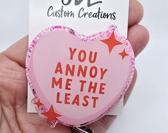May include: A pink heart-shaped badge reel with the text "YOU ANNOY ME THE LEAST" in red. The badge reel has a glittery border and red star accents. The top of the badge reel has a floral design and the text "JDL Custom Creations".
