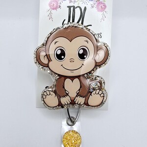 May include: A brown and white cartoon monkey with big eyes sits on a glitter-filled badge reel. The reel has a gold glitter circle attached to it.