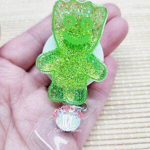 May include: Green glitter resin keychain with a cartoon character resembling a green gummy bear with a smiling face. The keychain is attached to a clear plastic badge holder with the text "JDL Custom Creations".