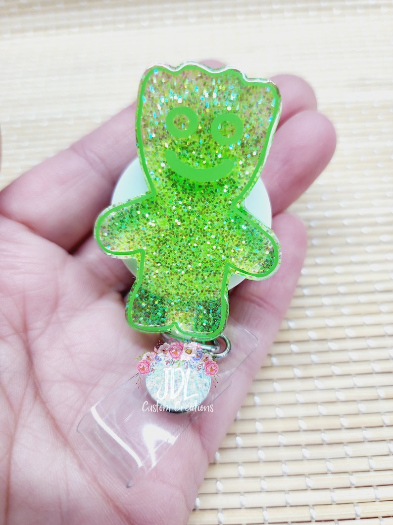 May include: Green glitter resin keychain with a cartoon character resembling a green gummy bear with a smiling face. The keychain is attached to a clear plastic badge holder with the text "JDL Custom Creations".