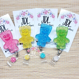 May include: Four glitter gummy bear badge reels in pink, yellow, blue, and green. Each reel has a clear plastic badge holder and a silver retractable cord.