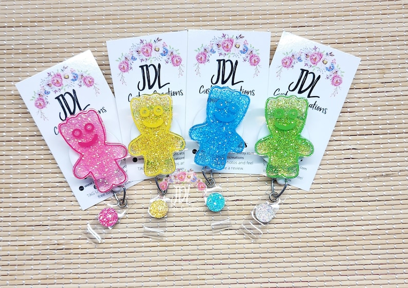 May include: Four glitter gummy bear badge reels in pink, yellow, blue, and green. Each reel has a clear plastic badge holder and a silver retractable cord.