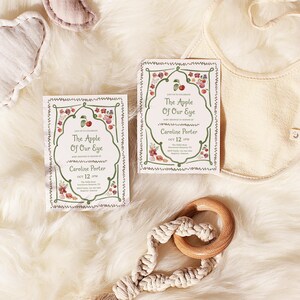 Puede incluir: Dos tarjetas rectangulares con un borde decorativo verde con ilustraciones de manzanas rojas y flores, texto central The Apple Of Our Eye, Caroline Porter y Oct 12 2018, junto a un babero color crema y un juguete de anillo de madera con macram&eacute;.