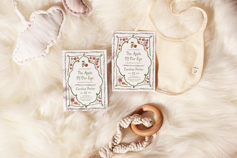 Puede incluir: Dos tarjetas rectangulares con un borde decorativo verde con ilustraciones de manzanas rojas y flores, texto central The Apple Of Our Eye, Caroline Porter y Oct 12 2018, junto a un babero color crema y un juguete de anillo de madera con macram&eacute;.