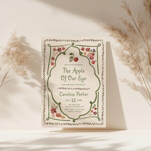Puede incluir: Una tarjeta de invitaci&oacute;n rectangular para baby shower con un borde verde dibujado a mano y bayas rojas. El texto dice The Apple Of Our Eye, en honor a Caroline Porter el 12 de octubre a las 2 PM en The Nellie Rose en Houston, Texas.