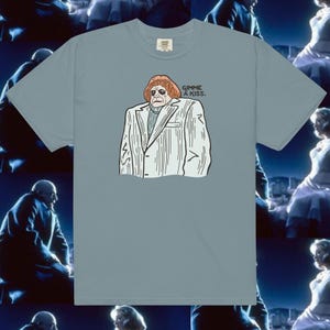 May include: A light blue t-shirt featuring a cartoon illustration of a person in a white suit with the text "Gimme a kiss." printed on the front.