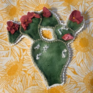 May include: A green fabric cactus applique with a white scalloped edge. The cactus features several dark red fabric flowers with small yellow centers attached to the top and sides. White thread is embroidered in small bird shapes on the surface.