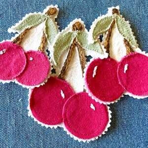 May include: Three fabric cherry patches featuring bright pink circular fruit, brown embroidered stems, and light green leaves. Each cherry pair is set against a white scalloped fabric backing with visible white stitching highlights on the fruit.