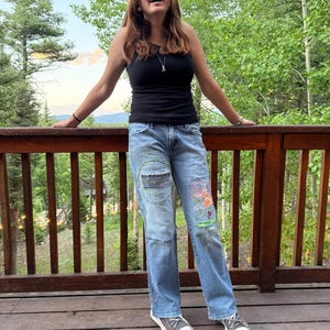 May include: A black tank top paired with light blue denim jeans featuring decorative embroidery, patches, and frayed edges. The jeans include a prominent circular green stitched design on the right thigh and colorful abstract patterns on the left leg.
