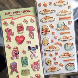 May include: Two sticker sheets. One features cartoon weightlifters with text including "Buff Puff Stickz" and "Don't Lose Your Whey." The other has egg-themed emojis with text like "Eggmoji Puffy Stickz!!" and "This is Fine."