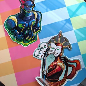 Puede incluir: Dos pegatinas con arte de fantasía. Una representa una figura humanoide azul con detalles amarillos y garras. La otra muestra una figura con un vestido rojo y dorado sosteniendo máscaras blancas con diversas expresiones.