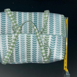 May include: A rectangular fabric bag featuring a repeating vertical pattern of green and teal leaves on a white background, with green and white gingham check handles. A yellow measuring tape shows the bag height is approximately 12 inches.