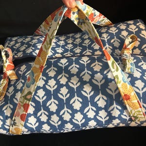 May include: A rectangular fabric bag featuring a blue and white floral pattern on the main body. The bag includes two handles and side ties decorated with a colorful floral print in shades of yellow, red, green, and blue against a white background.