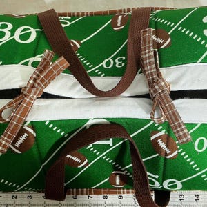 May include: A rectangular fabric storage basket with a green football field print featuring white yard lines and brown footballs. It has brown webbing handles, white trim, and brown plaid fabric ties. A ruler shows it is 17 inches wide.