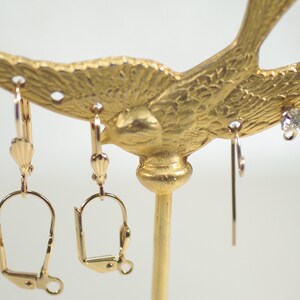 Pu&ograve; includere: Un espositore per orecchini color oro a forma di uccello. Diverse paia di orecchini sono appese all'espositore, tra cui orecchini a clip, a leva e a bottone. Gli orecchini presentano dettagli dorati e in cristallo trasparente.