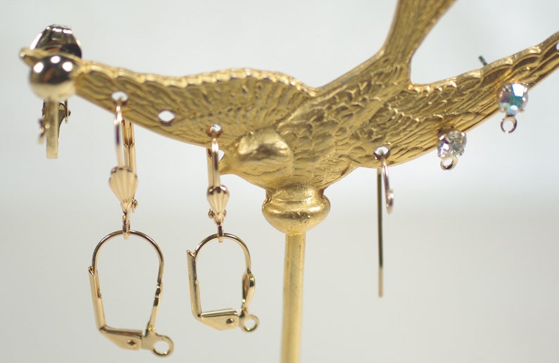 Pu&ograve; includere: Un espositore per orecchini color oro a forma di uccello. Diverse paia di orecchini sono appese all'espositore, tra cui orecchini a clip, a leva e a bottone. Gli orecchini presentano dettagli dorati e in cristallo trasparente.