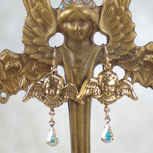 Pu&ograve; includere: Orecchini a forma di angelo dorati con un design a viso e ali di cherubino. Ogni orecchino presenta un pendente di cristallo iridescente a forma di goccia. Gli orecchini sono esposti su una statua di angelo dorata con un alone di cristalli iridescenti.