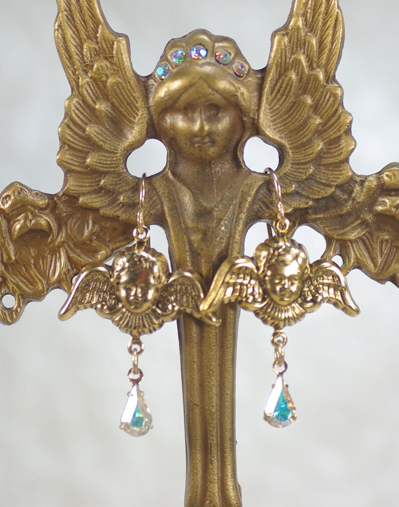 Pu&ograve; includere: Orecchini a forma di angelo dorati con un design a viso e ali di cherubino. Ogni orecchino presenta un pendente di cristallo iridescente a forma di goccia. Gli orecchini sono esposti su una statua di angelo dorata con un alone di cristalli iridescenti.