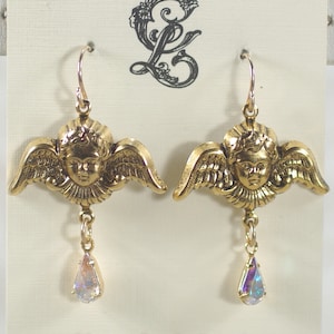 Pu&ograve; includere: Un paio di orecchini dorati a forma di angelo, con ali e volti dettagliati. Ogni orecchino ha un ciondolo iridescente a forma di goccia. Il testo "Classy Lady Jewelry" &egrave; stampato in basso.