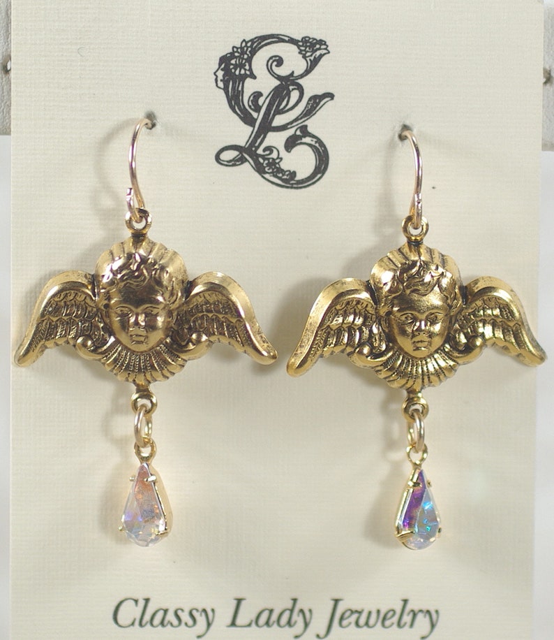 Pu&ograve; includere: Un paio di orecchini dorati a forma di angelo, con ali e volti dettagliati. Ogni orecchino ha un ciondolo iridescente a forma di goccia. Il testo "Classy Lady Jewelry" &egrave; stampato in basso.
