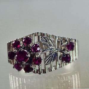May include: A silver-toned ring featuring a cluster of deep red gemstones and a stylized leaf design. The ring has a unique, openwork band with vertical bars. The gemstones are round and set closely together, creating a bold, eye-catching look.