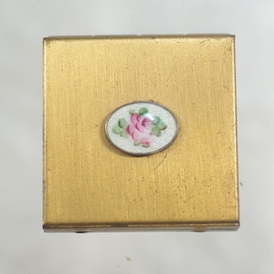 May include: A small, square, gold-toned metal compact. An oval, white enamel accent features a pink rose with green leaves. The compact has a slightly textured surface and a hinged design, suggesting it opens to reveal a mirror or storage compartment.