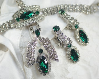 May include: A vintage jewelry set featuring a necklace and clip-on earrings. The set is silver-toned and adorned with clear rhinestones and emerald green oval and round gemstones. The jewelry is displayed on a white lace surface.