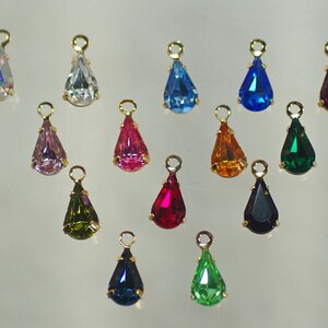 Pu&ograve; includere: Una collezione di ciondoli a forma di goccia in vari colori, tra cui blu, rosa, verde e viola. Ogni ciondolo ha un anello in metallo dorato nella parte superiore per appenderlo. I ciondoli sono esposti su uno sfondo chiaro.