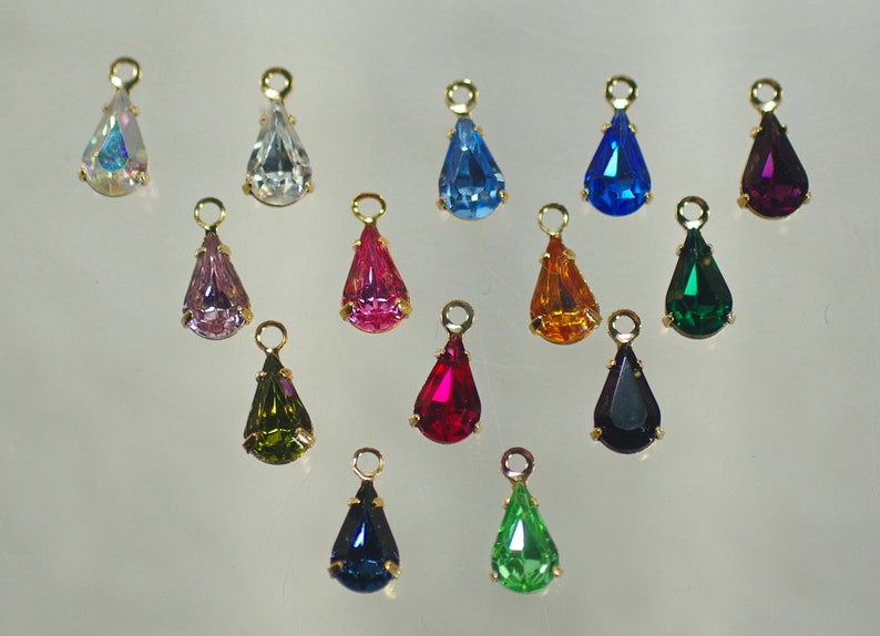 Pu&ograve; includere: Una collezione di ciondoli a forma di goccia in vari colori, tra cui blu, rosa, verde e viola. Ogni ciondolo ha un anello in metallo dorato nella parte superiore per appenderlo. I ciondoli sono esposti su uno sfondo chiaro.