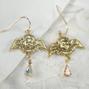 Pu&ograve; includere: Un paio di orecchini a forma di angelo color oro. Ogni orecchino presenta un viso di cherubino dettagliato con ali. Un cristallo iridescente a forma di goccia pende da ogni orecchino, aggiungendo un tocco di brillantezza. Gli orecchini hanno un classico design a gancio.