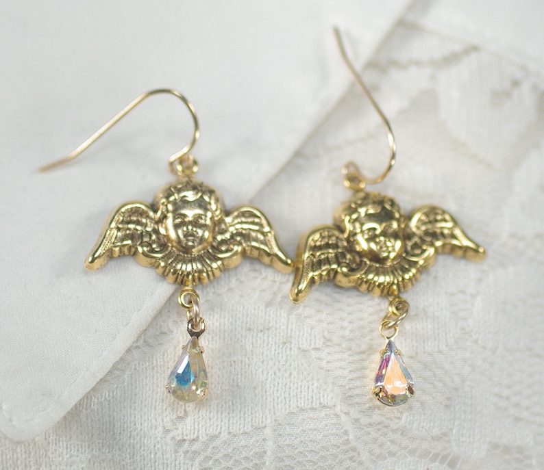 Pu&ograve; includere: Un paio di orecchini a forma di angelo color oro. Ogni orecchino presenta un viso di cherubino dettagliato con ali. Un cristallo iridescente a forma di goccia pende da ogni orecchino, aggiungendo un tocco di brillantezza. Gli orecchini hanno un classico design a gancio.