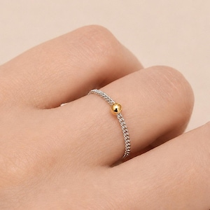 Könnte beinhalten: Ein dünner Ring aus einer silberfarbenen Kette mit einer einzelnen kleinen, polierten goldenen Perle in der Mitte. Der Ring besteht aus feinen, ineinandergreifenden silbernen Gliedern, die eine flexible Kreisform bilden und von der Kugel akzentuiert werden.