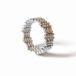 Puede incluir: Un anillo de tono plateado construido con múltiples filas de pequeñas cuentas esféricas pulidas. La banda presenta una superficie texturizada de cuentas con un acabado metálico reflectante, creando una forma circular uniforme que capta la luz.