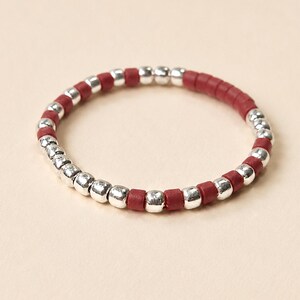 Peut inclure: Une bague circulaire composée d'une alternance de petites perles rondes argentées métalliques et de perles cylindriques rouge mat. Les perles sont enfilées selon un motif répétitif pour former un anneau continu et flexible d'épaisseur uniforme.