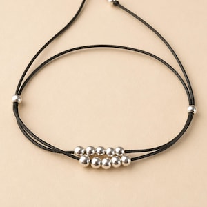 Peut inclure: Un bracelet en cordon noir composé de deux rangées de cinq petites perles sphériques argentées polies au centre, avec deux perles argentées supplémentaires placées sur les côtés et deux petites perles aux extrémités du cordon.