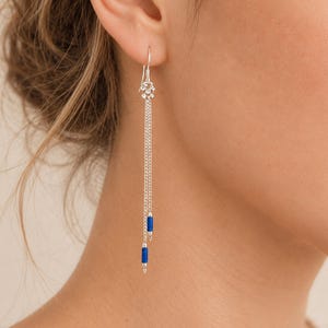 Peut inclure: Une boucle d'oreille pendante en argent composée d'un petit clou en forme de fleur relié à deux fines chaînes en argent de longueurs différentes. Chaque chaîne se termine par une petite perle cylindrique bleu profond, créant un design vertical.